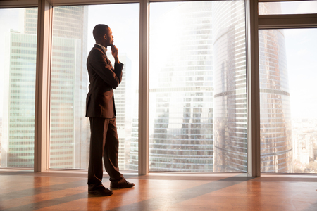 Young afro american contemplative businessman stands in office with hand on chin, looks through big window at the city, african entrepreneur enjoys sunset, rests after work, gets business inspirationの写真素材