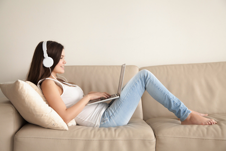 Attractive young woman in casual clothes and headphones lying on sofa and using laptop. Smiling teen girl listens and buying music in web store, watching online video, playing computer games at homeの写真素材