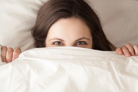 Young attractive woman with cunning look hiding face under white blanket at bedroom. Shy beautiful lady peeking from white duvet. Happy playful wife waiting husband in bed. Top view. Close up photoの写真素材