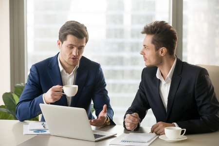 Two businessmen talking about new opportunities sitting with pc laptop at office desk, board of directors planning project, considering business offer, sharing ideas while drinking coffee togetherの写真素材