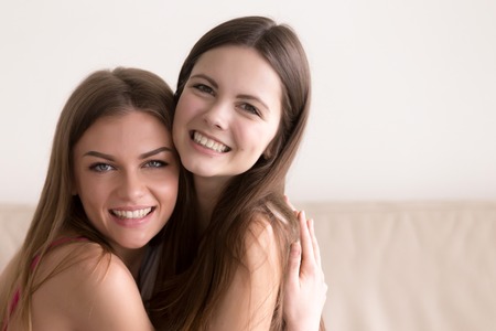 Close up headshot portrait of smiling young female friends warmly embracing during meeting. Two happy young women hugging and looking in camera. Strong friendship and sincere feelings between peopleの写真素材