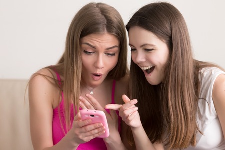 Close up portrait of two attractive young women sitting with cellphone, pointing on screen with surprise and joy. Girlfriends browsing funny photos from vacation, surfing dating web site, social mediaの写真素材