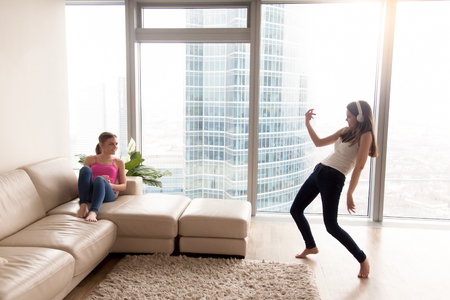 Pretty funny young woman enjoying music in stylish headphones, dancing in front of girlfriend sitting on sofa in bright living room. Female friends having fun while resting and fooling around at homeの写真素材
