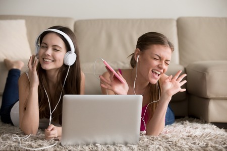 Two beautiful female music lovers in headphones enjoying popular musical compositions online with app on cellphone and laptop. Excited women friends singing songs and having fun while lying on floorの写真素材