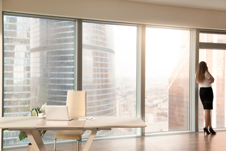 Modern office interior with desk and female silhouette standing back at full length window of business center, talking on phone, looking at cityscape, businesswoman having break at work after meetingの写真素材