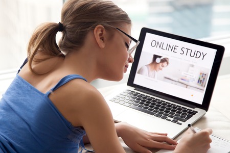 Shoulder view of young woman in eyeglasses writing with pen in paper notebook, laptop with online study landing page on screen on background. Female student searching courses, studying in Internet