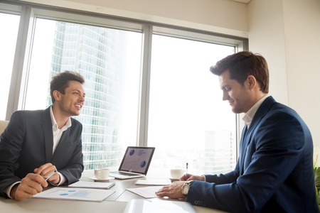 Satisfied businessman signing contract document while sitting at meeting desk in front of happy smiling business partner. Investor accepting agreement after studying project financial indicatorsの写真素材
