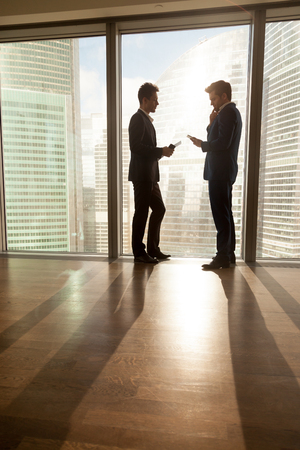 Business partners planning negotiation, exchanging contacts, thinking together about business development while standing near large window with modern urban landscape outside. Short meeting in officeの写真素材