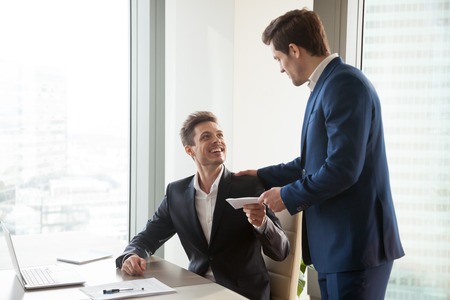 General manager presenting an envelope with premium or bonus cash to male company official. Boss congratulating happy employee with career promotion, thanking for good job and giving financial rewardの写真素材