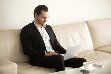 Handsome smiling young businessman looking at laptop screen. Project manager taking break from work, browsing internet, watching movie away from workplace. Successful entrepreneur relaxing on couch.の写真素材