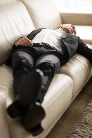 Tired exhausted businessman in suit laying on couch. Young hard working entrepreneur fell asleep on sofa at workplace. Staying at work overtime, overworked, too much workload, lack of sleep concept.の写真素材