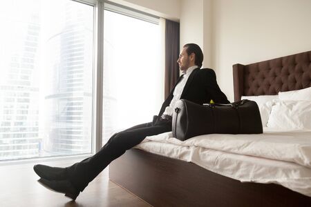 Relaxed businessman with luggage bag sitting on bed in luxurious hotel room, looking towards window. Smiling entrepreneur in suit rests after long business trip, looking forward to unpack and relax.の写真素材