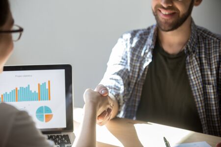 Two businesspeople shake hands to seal a deal with a smile, laptop with rising stats on screen on...