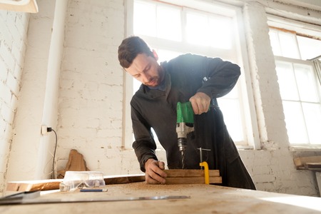 Skilled professional carpenter in workwear using electric drill machine, custom woodworking, making...