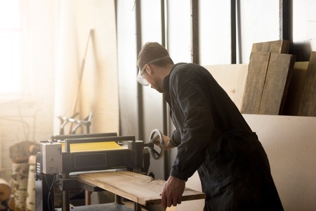 Back view of young carpenter or craftsman wearing working clothes and protective eyewear woodworking in small workshop interior, operating stationary power planer, processing trimming wooden plankの写真素材