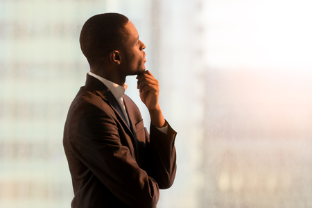 Portrait of pensive african american businessman standing near window and thinking about decision, dreaming of success, pondering new startup. Handsome black business leader imagining company futureの写真素材