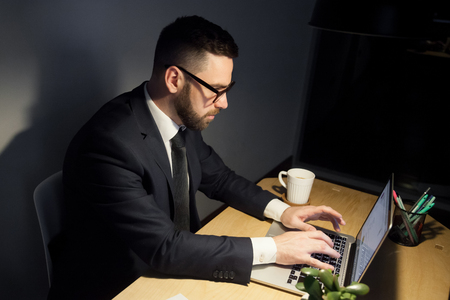 Attentive bearded man in glasses working on project late at night, typing messages, writhing emails using laptop computer. Forgotten cold cup of coffee or tea at his hand.の写真素材