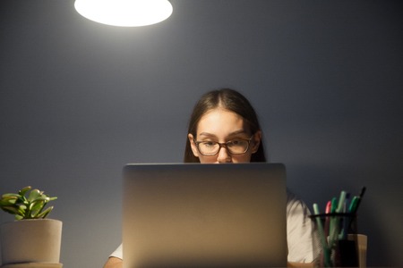 Overworked attentive young adult woman in glasses working on laptop computer at workplace desk late at night. Exam student preparing for test at home in dark room with lamp light. Front view.の写真素材