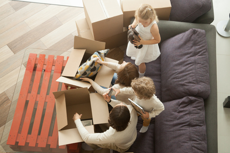 Kids helping parents to unpack boxes on sofa in cozy living room, happy young family with children having fun in new home together sitting on couch, moving into house relocation, top view from aboveの写真素材