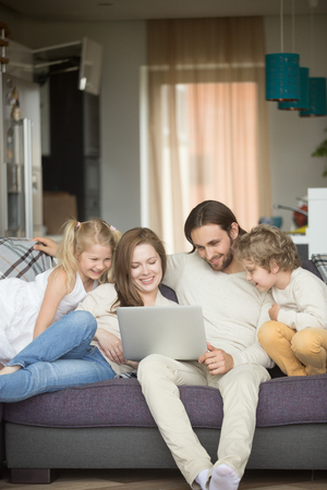 Family with children having fun using laptop sitting on sofa at home, parents and son daughter relaxing on couch holding computer, man woman with kids smiling watching funny video online, verticalの写真素材