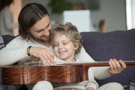 Young father teaching little son to play musical instrument at home, loving dad strumming guitar with small boy, parent and child relaxing on sofa, family leisure music activity, musically gifted kidの写真素材