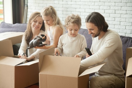 Happy family of four with two kids unpacking boxes moving into new home, smiling children helping parents with belongings in open cardboards sitting on sofa in living room together after relocationの写真素材