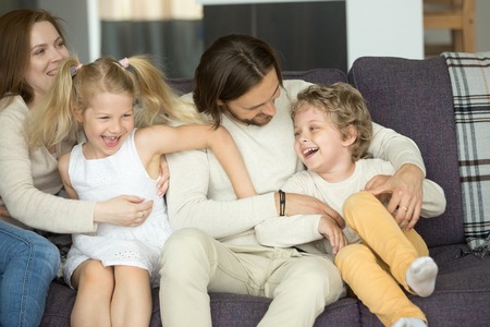 Happy parents and kids laughing having fun sitting on sofa, young married couple embracing preschool smiling son and daughter on couch, cozy warm loving caring family of four at home togetherの写真素材