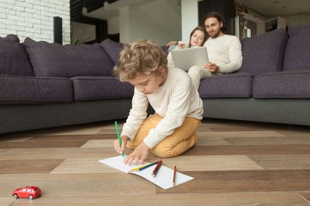 Preschool boy son drawing with colored pencils in album while parents using laptop on couch at background, creative little kid playing on living room floor, family leisure at home, child developmentの写真素材