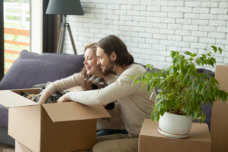 Young couple unpacking open cardboard boxes sitting on sofa in living room, happy new homeowners packing stuff moving out, smiling man and woman settle in new home, move and relocation conceptの写真素材