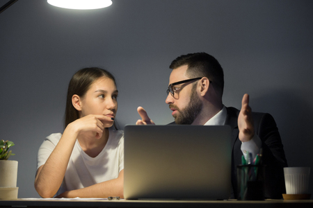 Bearded corporate manager in suit discussing ideas with millennial female worker at night shift. Business employers sitting at desktop behind laptop.の写真素材