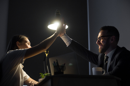 Millennial employers making high five at night shift in business office. Two happy successful young manager celebrating solution of job problem. Teamwork, winning, well done concept.の写真素材