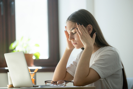 Exhausted young female worker is tired from work. Stress and headache behind laptop on job. Overwork, distress, negative emotions conceptの写真素材