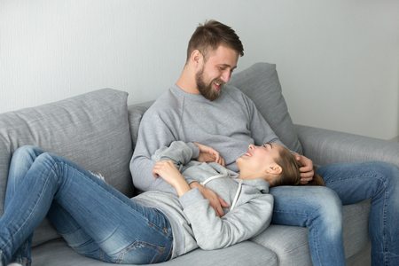 Loving affectionate young couple relaxing on couch together enjoying pleasant weekend at home, smiling man embracing stroking woman looking in the eyes, enjoying tenderness warmth in relationshipsの写真素材