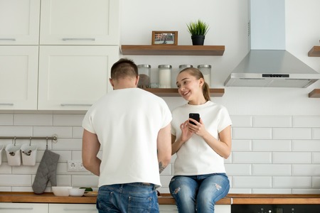 Young couple enjoying cooking in cozy kitchen, woman holding smartphone sitting on worktop talking to her man preparing food for breakfast, wife using mobile phone while husband making dinnerの写真素材