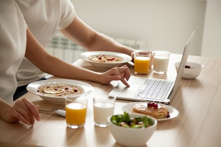 Couple having oatmeal and pancakes on breakfast using laptop on dining table, man and woman enjoying...