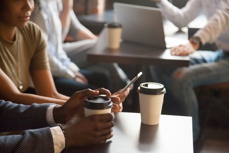 Coffee break in cafe with gadgets concept, diverse young african and caucasian people sitting at coffeehouse tables with drink to go in paper cups enjoy using mobile phone and laptop, close up viewの写真素材