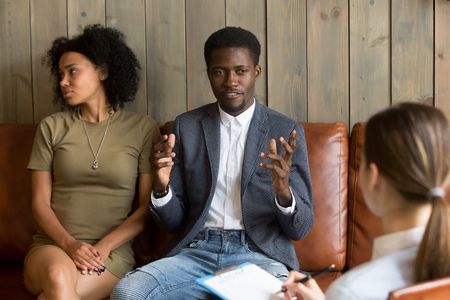 African man talking to family counselor, frustrated husband sharing marital problems while offended wife sitting silent on couch, black unhappy couple visiting psychologist, marriage counseling