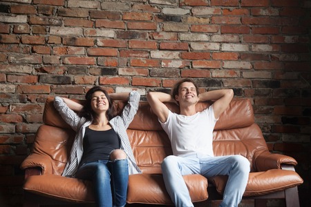 Young happy couple relaxing on comfortable leather couch at home, relaxed smiling man and woman resting on sofa in loft interior breathing fresh air meditating together, no stress, positive thinkingの写真素材