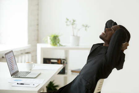 Satisfied relaxed african-american businessman in suit feeling happy at work sitting at office desk with laptop, smiling motivated black ceo enjoying good result dreaming of business success growthの写真素材