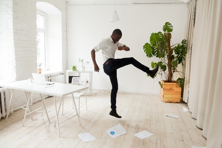 Emotional stressed african-american businessman fed up hates bad job kicking scattered papers on office floor, angry black employee quits going crazy fired on last day leaving work, nervous breakdownの写真素材