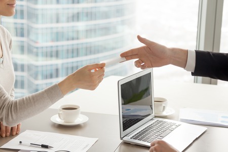 Businessman stretching out hand with blank business card over desk with laptop, coffee cups and documents to satisfied businesswoman after successful contract negotiation in company office. Close upの写真素材