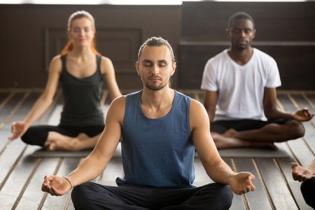 Group of young sporty afro american and caucasian people practicing yoga lesson with instructor, sitting in Sukhasana exercise Easy Seat pose, mudra gesture, students working out in club, indoorの写真素材