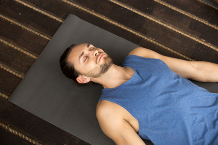 Young sporty man lying in Dead Body exercise or Corpse pose with his eyes closed, Savasana pose, working out, resting after practice, indoor close up, studio, top viewの写真素材