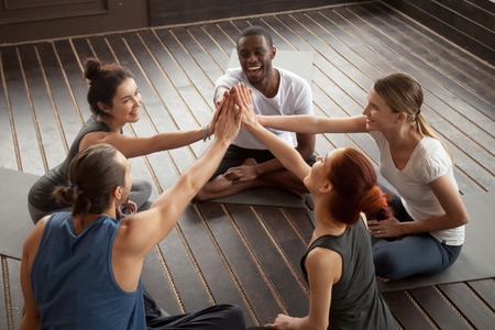 Diverse happy people giving high five sitting on mats at yoga seminar in studio, multiracial group joining hands celebrating good sport result, supporting motivation unity in fitness goal achievementの写真素材