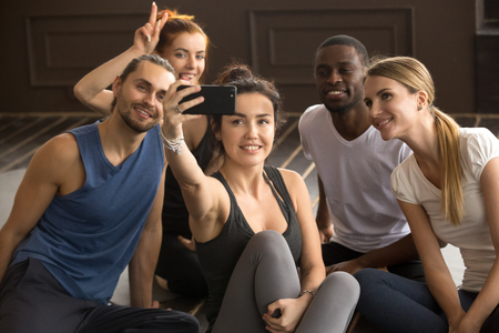 Sporty young fit smiling multi-ethnic people having fun taking group selfie in gym, diverse happy black and white friends making photo on smartphone together sitting in yoga studio at training breakの写真素材