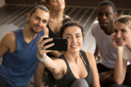 Taking selfie on smartphone concept, sporty woman holding cell shooting video or making diverse friends group self-portrait photo on modern smartphone camera at training break in gym, focus on phoneの写真素材
