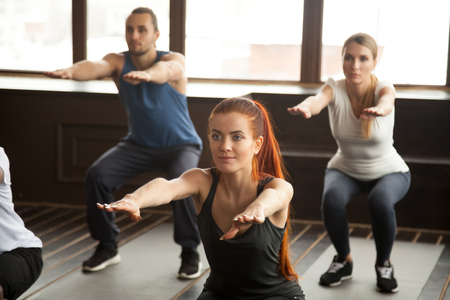 Sporty woman doing squat exercise at fitness training with young people, fit motivated group warming...