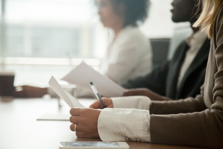 Diverse attentive people sitting at conference table holding documents at group meeting briefing, african and caucasian business seminar audience listening focused on training concept, close up view