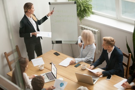 Businesswoman ceo boss in suit presenting corporate strategy pointing on flip chart at group meeting, business coach leader giving presentation explaining team goals in conference room at trainingの写真素材