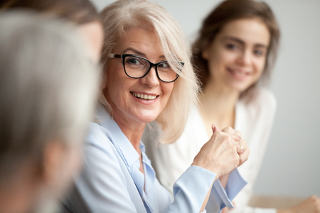 Smiling aged businesswoman in glasses looking at colleague at team meeting, happy attentive female team leader listening to new project idea, coach mentor teacher excited by interesting discussionの写真素材